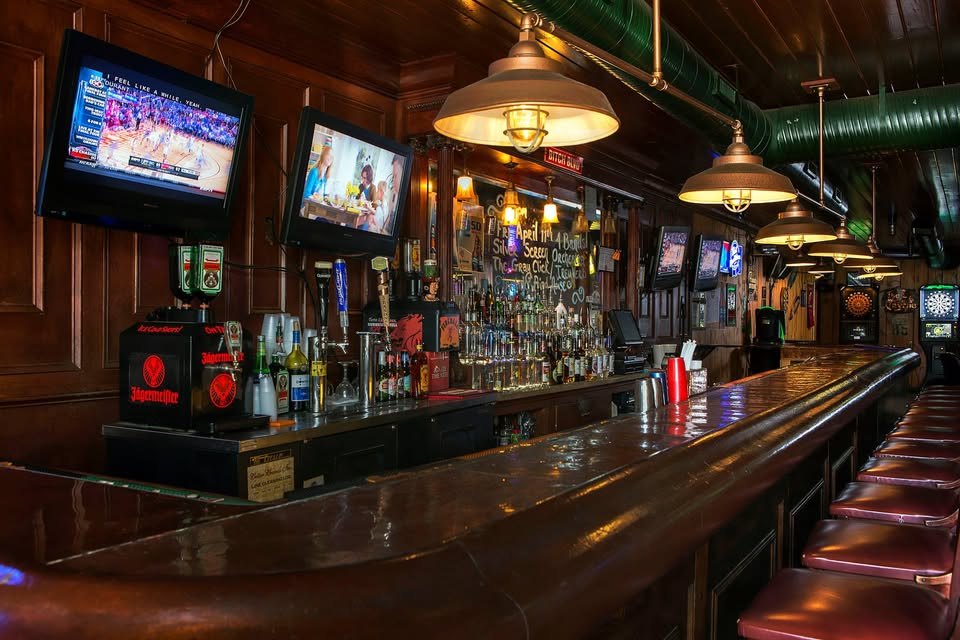 The long polished wood bar at The Tavrne, warm brass pendant lighting and a full liquor shelf